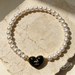 Puede incluir: Una pulsera de perlas con un dije en forma de corazón negro y un logotipo dorado. La pulsera está hecha de perlas blancas redondas. La joya se exhibe sobre una superficie clara.