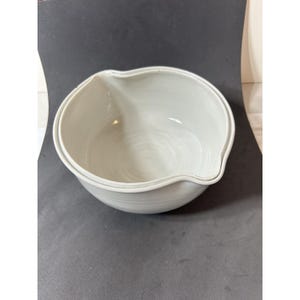 Może przedstawiać: Biała ceramiczna miska do mieszania z dzióbkiem. Misa jest okrągła, z szerokim brzegiem i gładkim, błyszczącym wykończeniem. Misa stoi na ciemnej powierzchni.