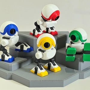 Peut inclure: Cinq petites figurines de robots blancs, avec des accents rouges, bleus, jaunes et verts. Chaque robot a une tête ronde et un grand œil circulaire. Ils sont disposés sur une plateforme grise à plusieurs niveaux.