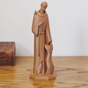 May include: A monochromatic brown figurine depicting a robed figure with a hood, standing on an octagonal base. The figure has a small bird on one shoulder, a dog looking up at the figure's hand, and a cat sitting near the base of the robe.
