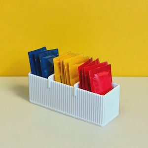 May include: A white, ribbed, three-compartment organizer filled with packets of various colors. The packets are blue, yellow, and red, and are arranged neatly within the organizer. The background is a solid yellow.