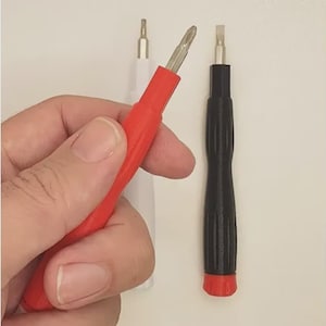 Puede incluir: Una mano sosteniendo un destornillador rojo con punta plateada, junto a un destornillador negro y un destornillador blanco. Los destornilladores tienen mangos texturizados sobre un fondo blanco.