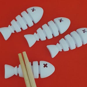 May include: White fish-shaped chopstick rests with a cross design on a red background. Two wooden chopsticks rest on one of the fish. The fish-shaped rests are a unique and decorative addition to any dining table.