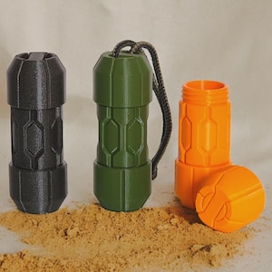 May include: Three cylindrical containers in black, green, and orange, with geometric designs. The green container has a black cord attached. The orange container is open, revealing the interior. The containers are on a sandy surface.