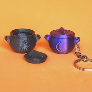May include: Two miniature cauldron keychains. One is black with a pentagram, the other is purple with a crescent moon. The keychains have handles and lids. The background is orange.