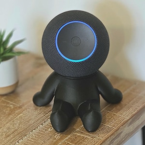 May include: A black spherical smart speaker with a textured mesh surface and a glowing blue circular light ring on top, mounted on a black figurine base shaped like a small seated character with outstretched arms and legs.