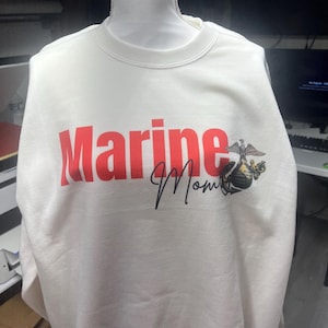 Könnte beinhalten: Weißes Sweatshirt mit dem Wort "Marine" in roten Blockbuchstaben und "Mom" in schwarzer Schrift. Auf der rechten Seite ist ein US Marine Corps-Emblem aufgedruckt. Das Sweatshirt wird auf einer Schaufensterpuppe präsentiert.