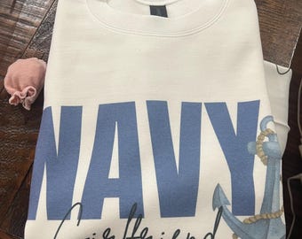May include: A white sweatshirt with the word "NAVY" in large blue letters and the word "Girlfriend" in a cursive font. An anchor graphic is on the right side. The sweatshirt is folded on a wooden surface.
