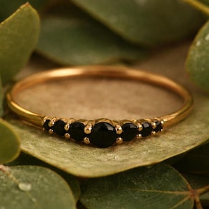 Peut inclure: Une bague en or ornée d'une série de pierres noires. La bague est posée sur un fond de feuilles vertes. Les pierres sont rondes et espacées de manière régulière, créant un design délicat et élégant. Un bijou.