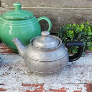 May include: Two teapots, one green and one silver, sit on a rustic wooden surface. The silver teapot has a black handle.
