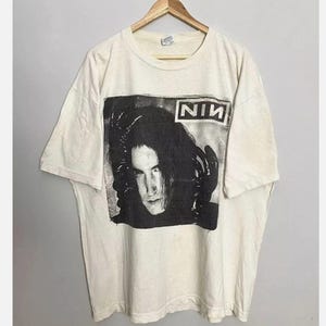 May include: Cream-colored t-shirt featuring a black and white image of a person with long hair and gloves. The word "NIN" is printed in a white box above the image. The shirt is hanging on a wooden hanger.