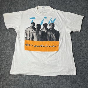 Könnte beinhalten: Weißes T-Shirt mit einer Grafik von vier Figuren in Schwarzweiß, mit dem Wort "Machine" in orangefarbener Schrift. Die Worte "T.M." stehen in Blau über den Figuren. Das Shirt hat einen Rundhalsausschnitt und kurze Ärmel.