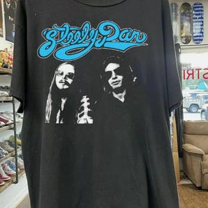 Puede incluir: Camiseta negra con el nombre de la banda "Steely Dan" en fuente azul claro. Debajo, una imagen en blanco y negro de dos hombres. La camiseta está colgada en una percha de madera.
