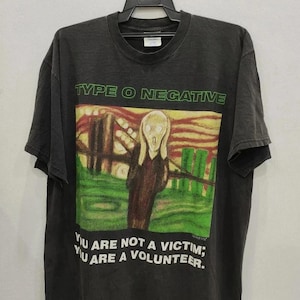 Może przedstawiać: Ciemnoszara koszulka z zielonym napisem "TYPE O NEGATIVE". Poniżej kolorowa grafika przedstawiająca postać w krajobrazie. Poniżej wydrukowano tekst "YOU ARE NOT A VICTIM; YOU ARE A VOLUNTEER".