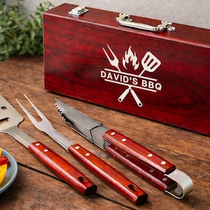 May include: A barbecue tool set consisting of a spatula, a two-pronged fork, and tongs, all featuring dark wood handles. The tools are displayed alongside a rectangular, dark red wooden storage case with a silver handle and the text DAVID'S BBQ.