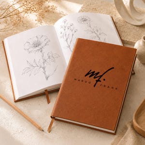 May include: A tan notebook with a brown cover featuring the black text "mf. MARCO FRANK" in a script and sans-serif font. The notebook is open to a page displaying a detailed black and white botanical line drawing of a flower with leaves.