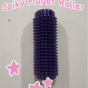 May include: A purple spiky fidget roller with a cylindrical shape. The roller has a textured surface with numerous small, pointed protrusions. The text "Spiky Fidget Roller" is above the roller. Pink star stickers are on the background.