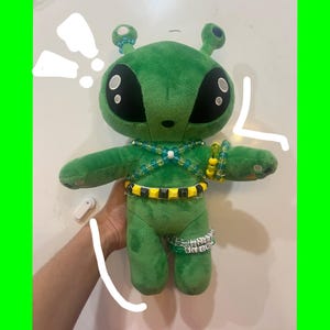 Puede incluir: Un muñeco de peluche verde con grandes ojos negros, antenas y un collar y pulseras de cuentas. El extraterrestre tiene un cinturón amarillo y negro y una pulsera en la pierna. El muñeco se sostiene sobre un fondo blanco.