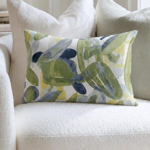 May include: A white, rounded armchair with three decorative cushions. The cushions include a rectangular cushion with a green and blue abstract design, a white cushion, and a light green cushion. The chair is near a window and a side table.
