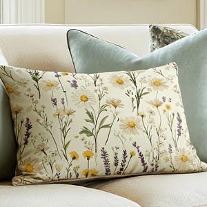 May include: A rectangular decorative pillow with a floral pattern featuring white daisies, yellow wildflowers, and purple lavender. Two light blue pillows flank the floral pillow on a cream-colored sofa. A framed picture hangs in the background.