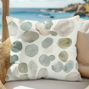 May include: Square decorative pillow with a cream background and watercolor-style abstract shapes in shades of green, gray, and beige. The pillow is placed on a wicker chair with a view of the ocean in the background.