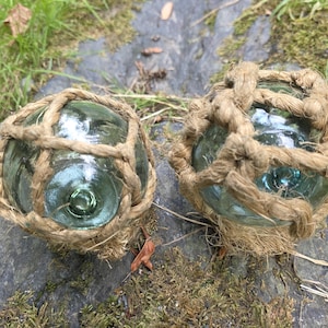 Op de afbeelding: Twee heldere glazen visdrijvers, elk omhuld met een geweven natuurlijk touwnet. De drijvers rusten op een rots met mos, met groen gras en een gevallen blad op de achtergrond. Het glas heeft een licht blauwgroene tint.