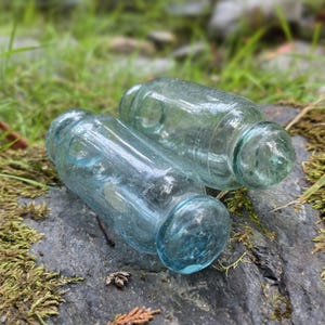 Könnte beinhalten: Zwei antike, klare Glasflaschen mit abgerundeten Böden und zylindrischen Körpern. Die Flaschen haben eine hellblau-grüne Farbe und liegen auf einem grauen Felsen mit grünem Moos und Gras im Hintergrund.