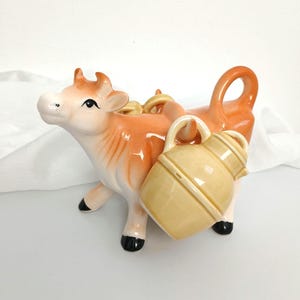 May include: A ceramic cow figurine with a light orange body, white face, and black hooves. The cow carries a light yellow jug with a handle. The figurine has a glossy finish and is set against a white background.