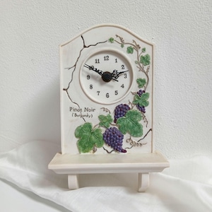 May include: Cream-colored decorative clock with a shelf base. The clock face has black hands and numbers. The clock is decorated with green leaves and purple grapes, and the text "Pinot Noir (Burgundy)" is visible.