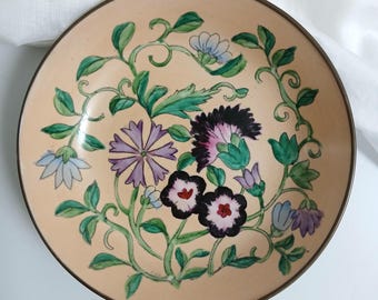 May include: A round ceramic plate with a light peach background featuring a hand-painted floral design. The pattern includes green vines, light blue, purple, and dark purple flowers with detailed petals and centers arranged in a circular motif.