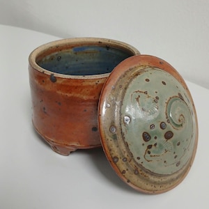 Puede incluir: Un tarro de cerámica hecho a mano con tapa. El tarro es cilíndrico con un exterior marrón rojizo y un interior azul. La tapa es de color verde claro con detalles marrones y blancos y un diseño en espiral.
