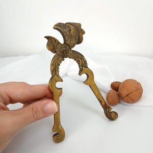 May include: A brass nutcracker featuring a detailed eagle head design at the top hinge. The tool has two long, curved handles with ornate leaf-like engravings along the sides and curled ends, showing a weathered, aged metallic finish.
