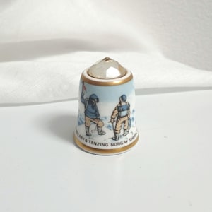 以下が含まれることがあります： 透明なファセット加工のトップと金色の縁が付いた磁器の指ぬき。指ぬきには、2人の登山者の絵が描かれており、ベースの周りには「Hillary & Tenzing Norgay Sherpa」という文字が書かれています。指ぬきは白で、青とタンのアクセントが付いています。
