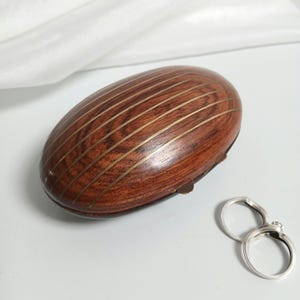May include: An oval-shaped wooden box featuring a dark brown wood grain with thin, parallel brass inlay stripes running across the top. Two simple silver-toned metal rings are positioned next to the box on a smooth, light-colored surface.