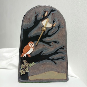 May include: A ceramic clock with a whimsical owl and tree design. The clock face features a brown owl perched on a black tree branch against a night sky backdrop. The clock hands are gold-colored. The clock measures approximately 6 inches tall.