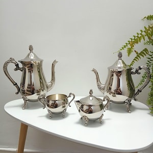 May include: A polished silver tea set comprising two teapots, a lidded sugar bowl, and a creamer. The set features decorative handles and feet. It is arranged on a white table with wooden legs, reflecting the room.