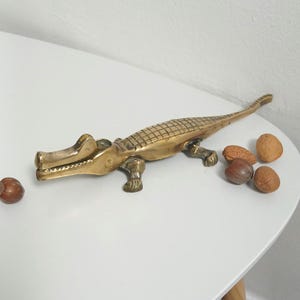 May include: A brass alligator nutcracker with a textured body and open jaws. Several nuts are scattered around the alligator on a white surface. The alligator is a decorative item and a functional tool.