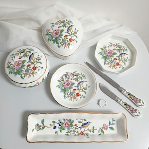 Op de afbeelding: Een verzameling wit keramisch serviesgoed met een bloemen- en vogelontwerp. Inclusief een hartvormige doos, een ronde doos, een rechthoekige dienblad, een kleine achthoekige schaal, twee messen en een kleine ronde schaal met ringen.