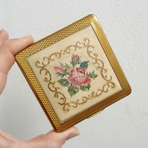 May include: A vintage gold-coloured compact with a square shape. The centre features a cream-coloured fabric with a cross-stitched floral design in pink, green, and blue. The compact has a textured border.