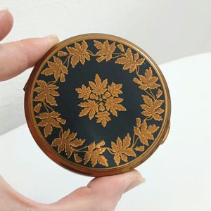 Peut inclure: Un poudrier rond vintage avec un dessus en émail bleu foncé. Le dessus présente un motif de feuilles de couleur or disposées en cercle autour d'un motif floral central. Le poudrier a un cadre en métal doré.