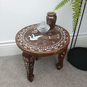 May include: A round, dark brown wooden side table with a peacock inlay design on the tabletop. The table has three curved legs and is decorated with white accents. A brown vase and a clear glass candle holder sit on top.