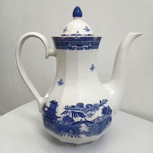 May include: White ceramic teapot with a blue and white landscape design. The teapot has a curved handle, a long spout, and a lid with a blue finial. The design includes boats, bridges, and buildings.