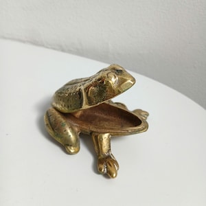 Könnte beinhalten: Eine goldfarbene Schmuckschatulle in Form eines Frosches, mit offenem Maul. Der Frosch hat eine strukturierte Oberfläche und detaillierte Merkmale. Die Box steht auf einer weißen Oberfläche.