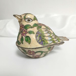 以下が含まれることがあります： 鳥の形をした装飾的な小物入れ。ボックスはクリーム色で、ピンク、緑、紫の花柄と葉柄のデザインが施されています。鳥は金色のくちばしとトリムを持っています。