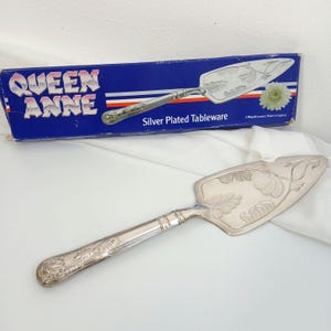 May include: Silver-plated cake server with an ornate handle and a blade decorated with leaf patterns. The server is displayed with its original blue box, which reads "Queen Anne Silver Plated Tableware."