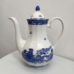 May include: White ceramic teapot with a blue and white landscape design. The teapot has a long spout, a curved handle, and a lid with a blue finial. The design features a pagoda, trees, and a bridge.