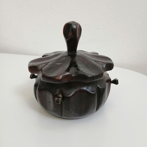 Op de afbeelding: Een donkerbruine houten sieradendoos met een bloemvormig deksel en een gebogen handvat. De afgeronde doos heeft kleine metalen sluitingen. De doos staat op een wit oppervlak. De doos is waarschijnlijk van donker hout.