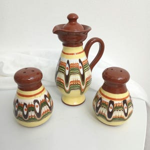 May include: A ceramic condiment set consisting of a lidded pitcher and two matching shakers. Each piece features a cream-colored base with a brown top and a decorative pattern of wavy, marbled lines in shades of dark brown, green, and orange.