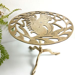 May include: A brass trivet with a squirrel design. The round trivet has a three-legged base and a decorative cutout pattern featuring a squirrel holding a nut. The brass has a warm, golden hue, and the design is detailed and intricate.