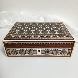 May include: A rectangular wooden box featuring intricate geometric marquetry patterns on the lid and front panel. The design consists of small triangular inlays in shades of brown, white, and dark wood, with a central keyhole on the front face.
